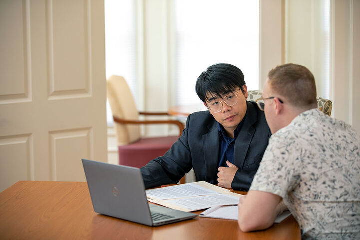 Two men with a laptop and papers sit at a table and have a discussion.